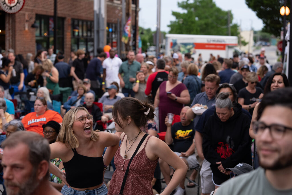 Large group of people enjoying a performance. Image is focused on two girls laughing to one another in the center.